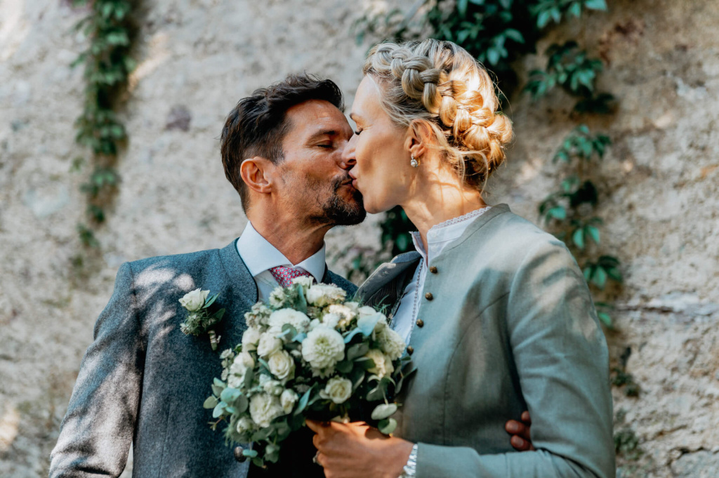 Brautpaar küsst sich liebevoll vor einer mit Efeu bewachsenen Wand.