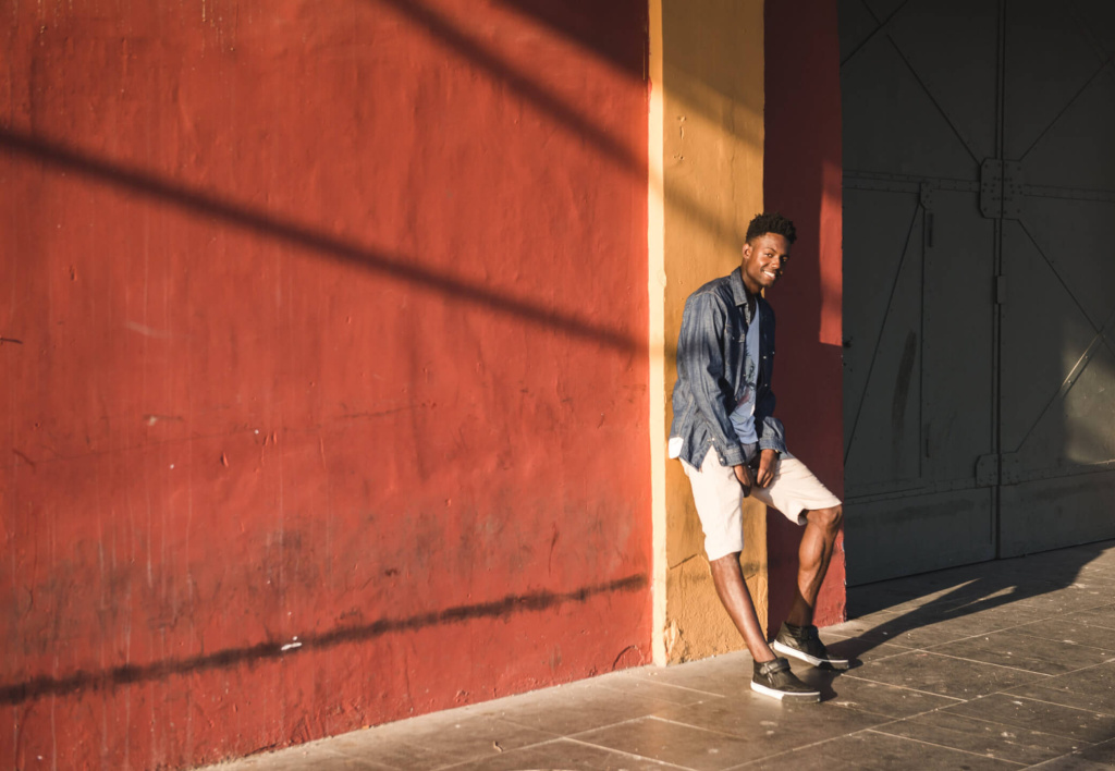 Junger Mann in legerer Kleidung lehnt an einer bunten Wand in warmem Licht.